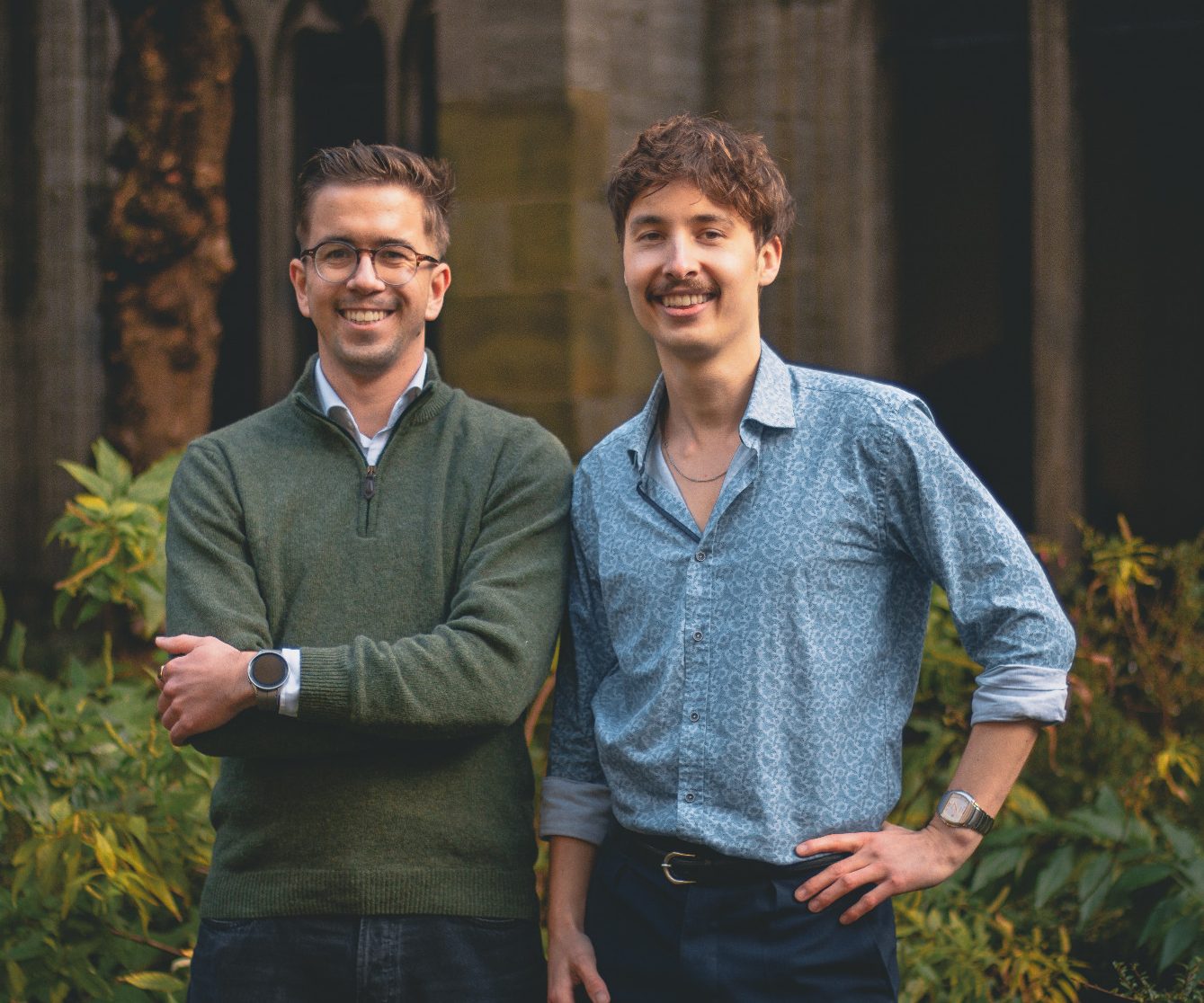 Twee mannen met glimlachen, staand in een groene omgeving met planten op de achtergrond.